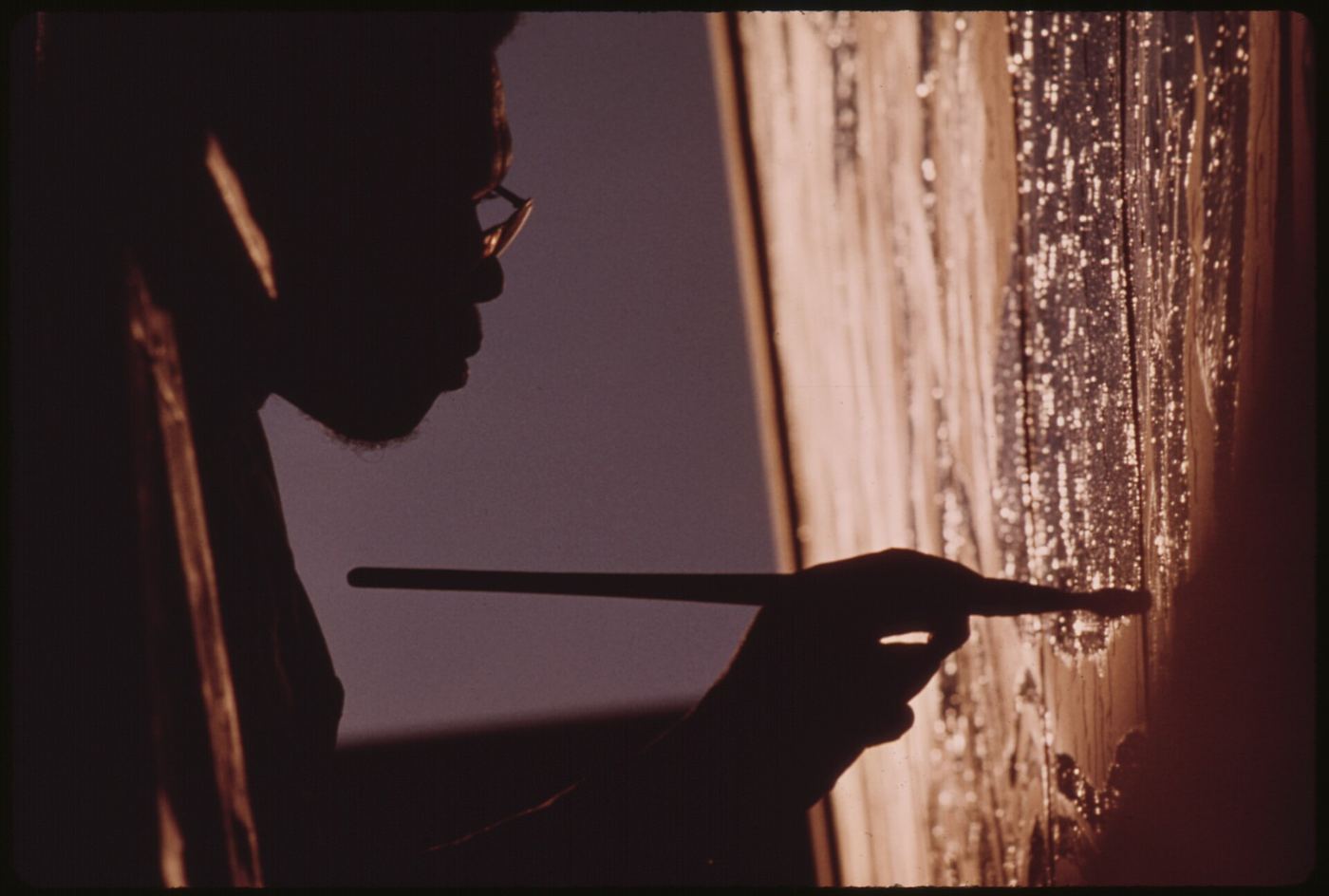 Silhouette of an artist's hand drawing with a long brush against a lit wall